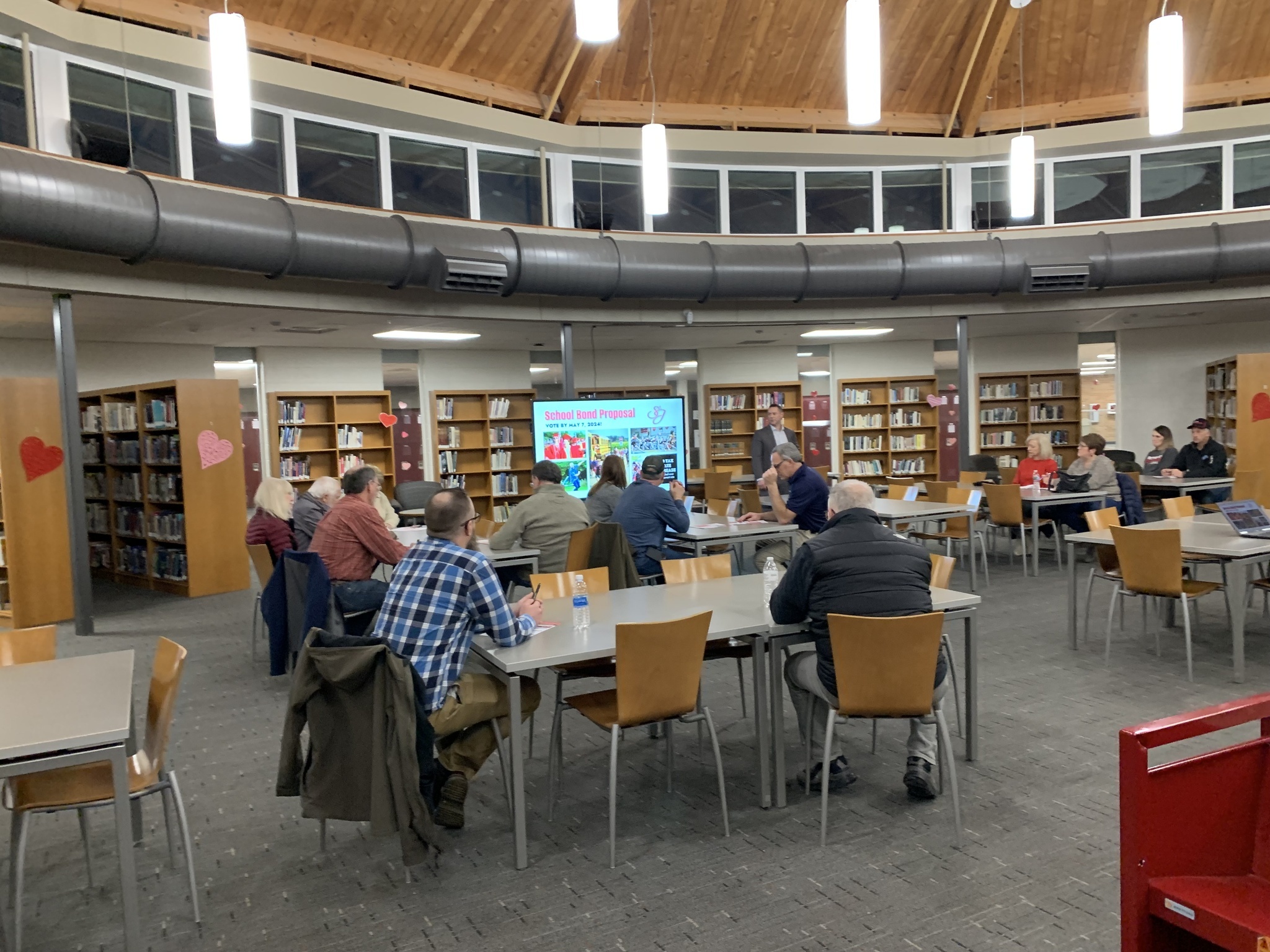 A group of community members gathers in the high school media center as Superintendent Dr. Anthony Berthiaume presents a slide titled "2024 School Bond Proposal"